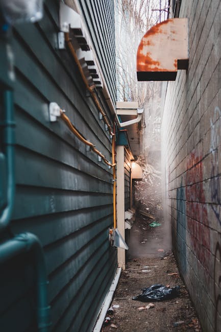 A narrow alleyway between two buildings, with the left featuring dark green horizontal siding and visible utility pipes and conduits attached along its surface. The right side displays a weathered brick wall with some faint graffiti markings. In the background, a partially visible outdoor space shows leafless trees and an overcast sky. The ground is uneven, with patches of dirt, scattered rubbish, and a plastic bag near the base of the brick wall, indicating a typical alley used for waste collection or clearance. An outdoor wall-mounted light fixture provides minimal illumination, while a large, rusty, semi-circular metal cover extends from the brick wall, possibly for ventilation or drainage purposes. This scene exemplifies an environment where independent waste removal services, such as those provided by [COMPANY_NAME], might operate to manage rubbish in private or less accessible areas, supporting alternative waste handling outside standard municipal collection routes.