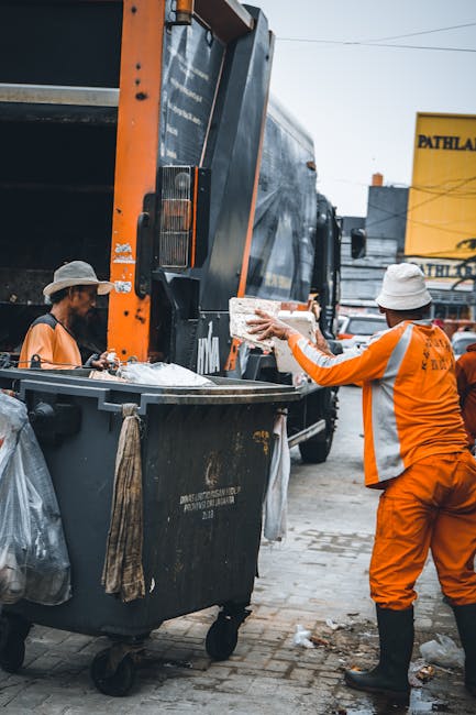 In an outdoor urban setting, two waste collection workers are engaged in rubbish removal near a large black industrial waste bin with a metal lid. The worker on the right, dressed in bright orange high-visibility clothing, a white hat, and black boots, is placing a piece of debris into the bin. The other worker, also in orange high-visibility gear and a beige hat, stands beside the bin, managing waste. Behind them, a large black refuse collection truck with orange accents is parked, with its rear compartment open, ready to empty the bin. The ground is paved with concrete tiles, with some scattered rubbish and debris. In the background, there are commercial buildings, including a sign that reads 'PATHWAY,' and utility wires are visible in the sky. The scene illustrates typical private waste collection activities associated with on-site rubbish clearance and alternative waste handling services, as provided by companies like rubbishcollectionkentishtown.co.uk, aimed at managing municipal and construction waste efficiently in an urban environment.