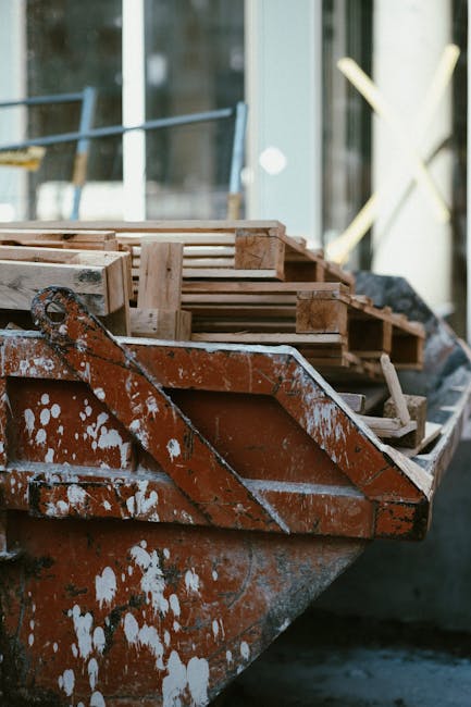 A close-up view of an old, weathered metal boat with visible rust and peeling white paint, docked alongside a construction site or storage area. Inside the boat, several wooden pallets are stacked, made of light-colored timber with a smooth, unfinished surface. The pallets are arranged in a slightly uneven manner, with some edges protruding over the sides of the boat. In the background, blurred elements of a building with glass windows and metal railings suggest a modern urban environment, possibly near a property undergoing refurbishment or cleanup. Natural daylight illuminates the scene, highlighting the textures of the rusted metal surface and the wooden pallets, creating a professional and neutral tone suitable for describing waste management or rubbish removal services, such as those provided by [COMPANY_NAME], focused on alternative waste handling methods. The setting indicates an area where construction debris or discarded materials, including wooden pallets and metal components, are temporarily stored before disposal or recycling as part of a private rubbish collection effort near Kentish Town.