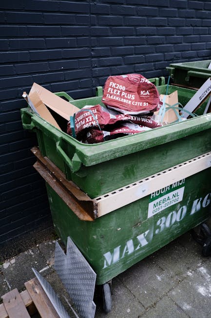 In an outdoor urban setting, two waste collection workers are engaged in rubbish removal near a large black industrial waste bin with a metal lid. The worker on the right, dressed in bright orange high-visibility clothing, a white hat, and black boots, is placing a piece of debris into the bin. The other worker, also in orange high-visibility gear and a beige hat, stands beside the bin, managing waste. Behind them, a large black refuse collection truck with orange accents is parked, with its rear compartment open, ready to empty the bin. The ground is paved with concrete tiles, with some scattered rubbish and debris. In the background, there are commercial buildings, including a sign that reads 'PATHWAY,' and utility wires are visible in the sky. The scene illustrates typical private waste collection activities associated with on-site rubbish clearance and alternative waste handling services, as provided by companies like rubbishcollectionkentishtown.co.uk, aimed at managing municipal and construction waste efficiently in an urban environment.