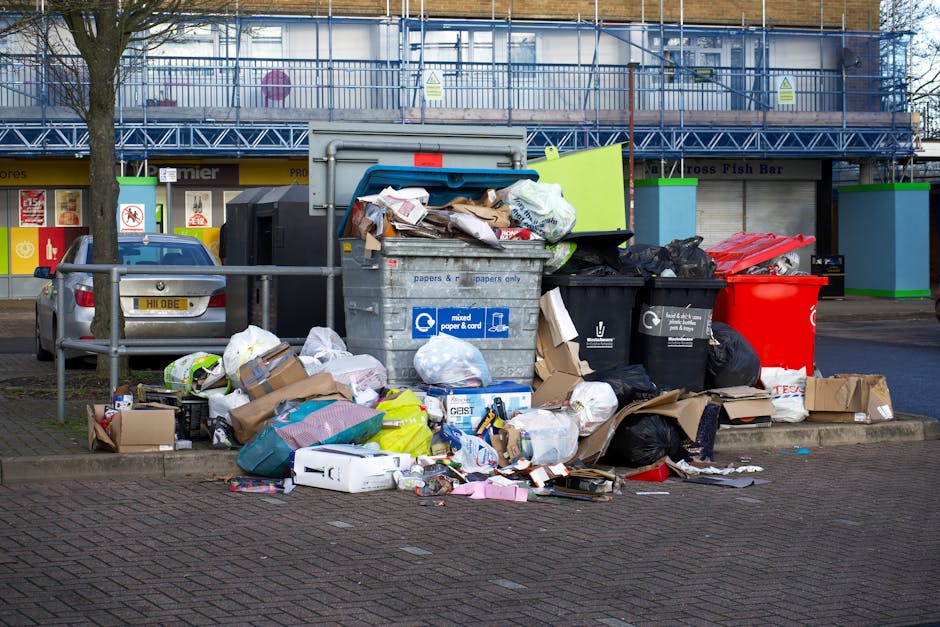 A large outdoor waste collection point situated on a paved street, featuring multiple black and red recycling bins filled with various types of refuse. The grey bin labeled for mixed paper and cardboard is overflowing with crumpled newspapers, flattened boxes, and other paper packaging spilling onto the ground. Surrounding it are several plastic bags, some black and others white, containing additional waste materials, alongside neatly flattened cardboard boxes. A small silver car is parked close by, partially obstructing a metal railing in front of the bins. In the background, there is a commercial building with storefronts, including a visible sign for Fish Bar and other shops, and a blue sculpture or sign attached to the railing. The scene takes place during daylight hours under an overcast sky, and the overall environment appears to be a designated rubbish disposal area reflecting the need for proper waste management in accordance with local rules for rubbish disposal, possibly facilitated by independent waste collection services like those offered by Rubbish Collection Kentish Town.