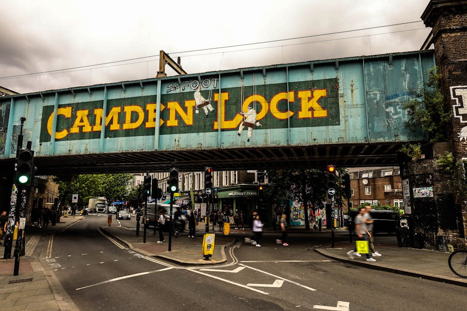 A weathered railway bridge painted in a faded turquoise hue with rust spots and peeling paint, prominently displaying the words 'Camden Lock' in large yellow letters, spans across a busy urban street scene. The bridge features graffiti and murals, including illustrations of children hanging from the letters. Below the bridge, a variety of pedestrians are walking on the pavement, some carrying bags, while others are waiting to cross at multiple traffic lights painted with green signals. A black van and several cars are visible on the road, with road markings and bicycle lanes clearly demarcated. A yellow emergency cone is positioned near the curb, and in the background, there are residential buildings, shops, and street art on walls. The ambient lighting is dull with overcast skies, creating a muted atmosphere. This scene exemplifies urban infrastructure and street activity, which some private waste management services like rubbish collection companies in Kentish Town might attend to for on-site clearance or alternative waste handling when managing rubbish removal in such high-traffic city areas.
