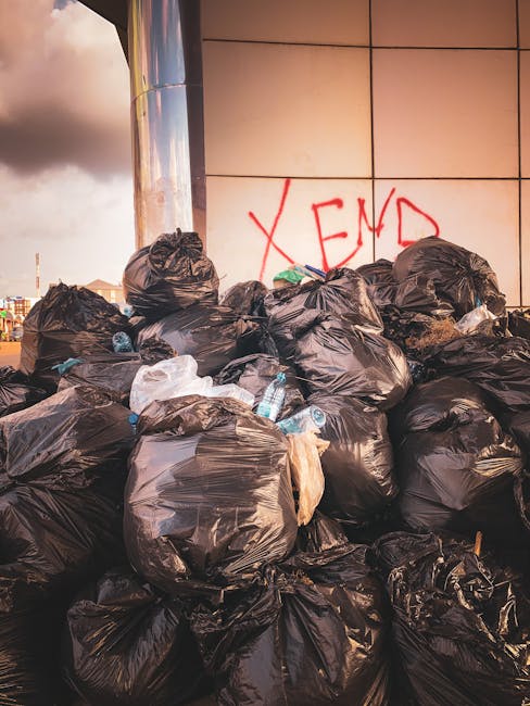 A large outdoor waste collection point situated on a paved street, featuring multiple black and red recycling bins filled with various types of refuse. The grey bin labeled for mixed paper and cardboard is overflowing with crumpled newspapers, flattened boxes, and other paper packaging spilling onto the ground. Surrounding it are several plastic bags, some black and others white, containing additional waste materials, alongside neatly flattened cardboard boxes. A small silver car is parked close by, partially obstructing a metal railing in front of the bins. In the background, there is a commercial building with storefronts, including a visible sign for Fish Bar and other shops, and a blue sculpture or sign attached to the railing. The scene takes place during daylight hours under an overcast sky, and the overall environment appears to be a designated rubbish disposal area reflecting the need for proper waste management in accordance with local rules for rubbish disposal, possibly facilitated by independent waste collection services like those offered by Rubbish Collection Kentish Town.