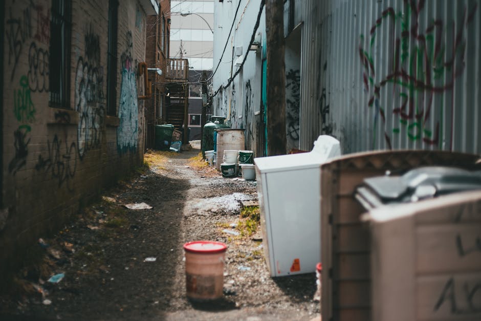 The image depicts a narrow alleyway situated between two urban buildings, with one side featuring an exposed brick wall covered in multicoloured graffiti, and the other side a corrugated metal fence also marked with spray paint. The alley's uneven, muddy ground is strewn with a few scattered pieces of litter. Various discarded household items and waste containers are lined along the pathway, including a white plastic washing machine or appliance, a large brown and beige trash bin, and several green and white bins placed near the far end. Some of these containers and objects are leaning, stacked, or positioned randomly, indicating an informal rubbish collection or abandoned waste. In the background, there are additional waste bins and intermittent wooden structures or staircases leading to upper levels of nearby buildings, contributing to an urban, slightly neglected atmosphere. The lighting suggests daylight, with natural light illuminating the scene subtly, emphasizing the textures of the trash and graffiti, and subtly relating to alternative waste handling or informal rubbish accumulation in tight city spaces.