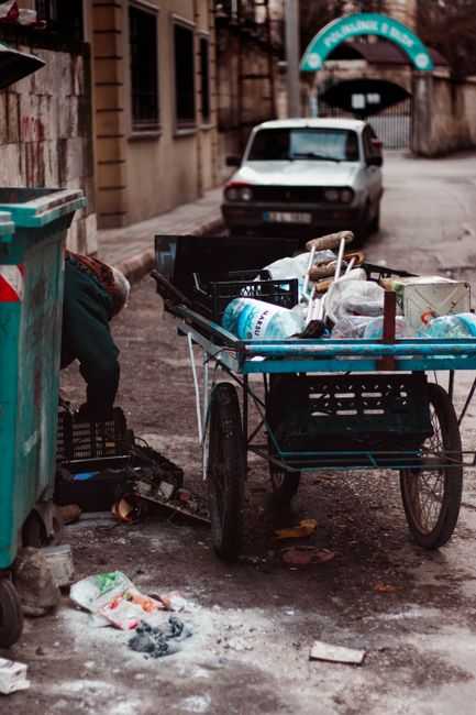 The image depicts a narrow alleyway situated between two urban buildings, with one side featuring an exposed brick wall covered in multicoloured graffiti, and the other side a corrugated metal fence also marked with spray paint. The alley's uneven, muddy ground is strewn with a few scattered pieces of litter. Various discarded household items and waste containers are lined along the pathway, including a white plastic washing machine or appliance, a large brown and beige trash bin, and several green and white bins placed near the far end. Some of these containers and objects are leaning, stacked, or positioned randomly, indicating an informal rubbish collection or abandoned waste. In the background, there are additional waste bins and intermittent wooden structures or staircases leading to upper levels of nearby buildings, contributing to an urban, slightly neglected atmosphere. The lighting suggests daylight, with natural light illuminating the scene subtly, emphasizing the textures of the trash and graffiti, and subtly relating to alternative waste handling or informal rubbish accumulation in tight city spaces.