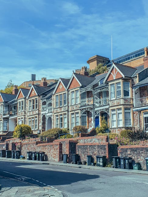 A row of Victorian-style terraced houses with decorative white wrought iron balconies and large bay windows facing a paved street. The houses have brick facades in shades of red and brown, with some featuring ornate woodwork detailing around the windows and gabled roofs. The front gardens are raised and enclosed by low brick walls, with neatly trimmed bushes and small trees. Along the sidewalk, several black waste bins are lined up in front of the properties. Behind the row of houses, there is a modern building with large glass windows and a rooftop terrace, indicating an urban residential area. The sky is clear with a few wispy clouds, and sunlight illuminates the scene, highlighting the textures of the brickwork and architectural details. This setting reflects a typical residential street where private waste collections or alternative rubbish removal services like those provided by Kentish Town waste specialists could operate to manage household waste efficiently.