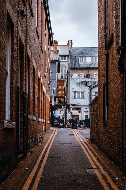 A narrow alleyway situated between two brick buildings in an urban area, with the alley paved in asphalt featuring double yellow lines along its edges. The left and right sides are lined with red and brown brick walls, each with several small, rectangular windows, some with white window frames. In the background, multi-story residential buildings with grey and weathered facades are visible, including rooftop structures, vents, and chimneys. A metal staircase with wooden steps is attached to the back of one building, leading upwards. The alley appears clear of rubbish but is a typical setting where excess waste, such as discarded furniture or packaging, might be temporarily stored before collection. The sky above is overcast, casting a diffuse, natural light over the scene, emphasizing textures of the brickwork and pavement. This setting reflects common urban environments where private waste collection or rubbish removal services might operate, such as those offered by Kentish Town waste management providers.