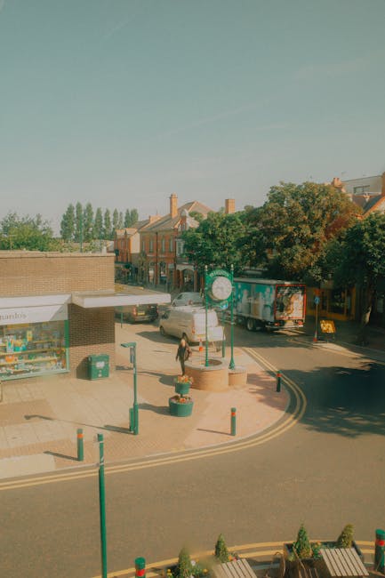 The image shows a small urban street scene during daylight hours, with a pedestrian walking along the sidewalk near a row of parked vehicles, including a white van and a truck. In the foreground, there are decorative flower pots and green bollards along the edge of the paved area, which features a curved road with striped double yellow lines indicating it is a main thoroughfare. To the left, a single-story building with a shopfront displays various items behind a large window, and a trash bin is positioned nearby, indicating urban waste management practices. Behind the sidewalk, there is a line of mature trees with dense foliage, providing shade and a natural backdrop to the residential and commercial buildings, visible in the background with brick facades and chimneys. The overall scene appears quiet and well-maintained, with natural lighting and shadows cast by nearby trees and vehicles, aligning with private waste collection themes by illustrating an organized street environment where rubbish removal services such as those by [COMPANY_NAME] might operate or support alternative waste handling arrangements in the local area.