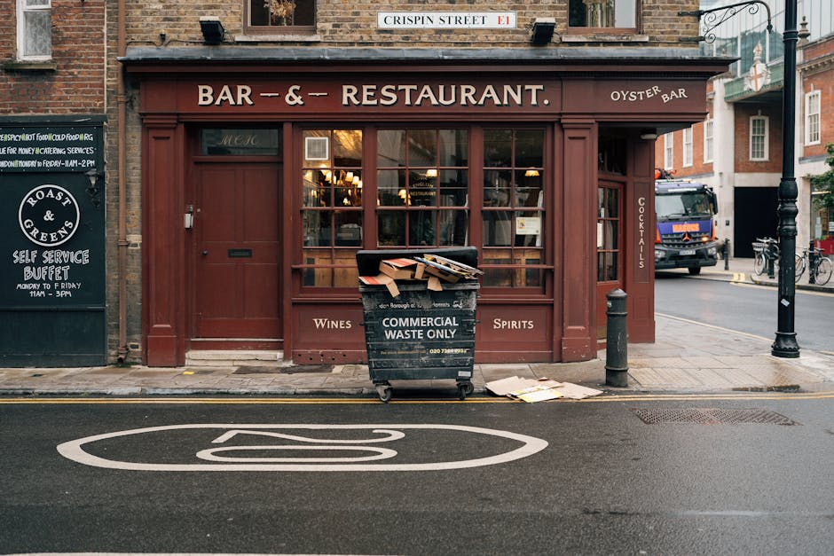 In front of a red-brick building housing a bar and restaurant, a black commercial waste bin marked 'COMMERCIAL WASTE ONLY' is placed on the sidewalk, filled with scattered cardboard boxes and paper. The bin is positioned directly outside a large window with a wooden frame, and the storefront displays signage indicating offerings such as wines, spirits, and cocktails. To the left, a black sandwich board advertises a self-service buffet, while a street sign reading 'CRISPIN STREET E1' is mounted above the establishment. Further to the right, a parked dark-blue truck is partially visible on the street, along with a metal lamppost and bicycles secured to a rack. The scene captures an urban area with a paved road featuring a white line and a piece of artwork painted on the pavement, suggesting nearby vehicle or bicycle parking. This setting relates to rubbish removal services that handle commercial waste collection outside dining or hospitality venues, exemplifying the typical use of private waste disposal solutions managed by firms like Rubbish Collection Kentish Town, in contrast to local authority bin collections.