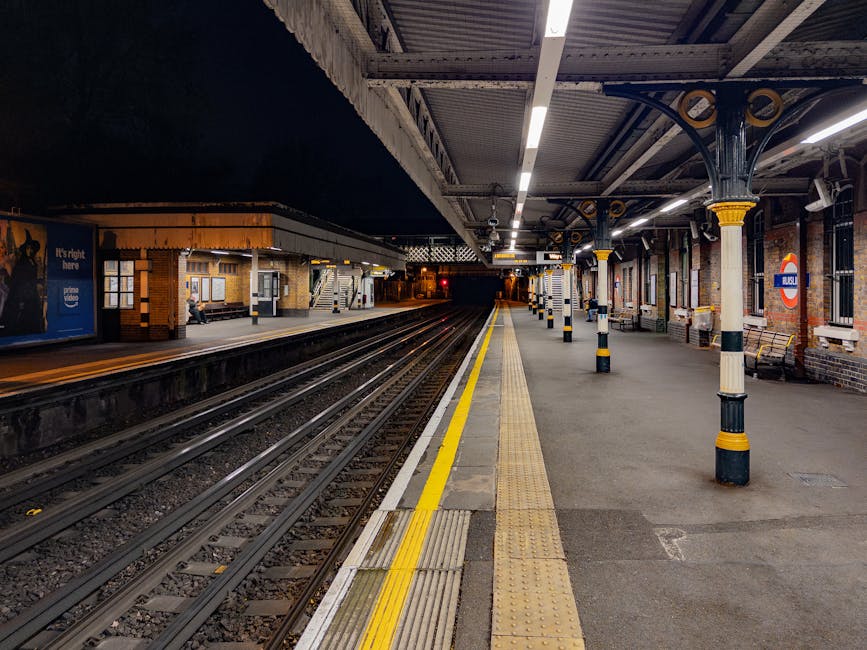 The image depicts an empty train station platform at night, illuminated by overhead lighting. The platform has a textured yellow safety line and tactile paving along its edge, with a smooth asphalt surface adjacent to the railway tracks. Multiple black-and-white striped bollards are positioned along the platform, close to the edge, separating waiting areas from the trackside. The station features brick walls with several windows, some of which have posters or advertisements, and there is a sign indicating the station's name as part of the London Underground network. In the background, a small shelter with a bench and a person sitting nearby is visible, with additional platform sections extending into the darkness. The scene is well-lit, with the platform's ceiling supported by ornate metal columns with decorative yellow accents. The environment is clean and organized, reflecting a typical urban train station prepared for passenger use and potential adoption of private waste handling or cleaning services by the station management, akin to independent rubbish collection services.