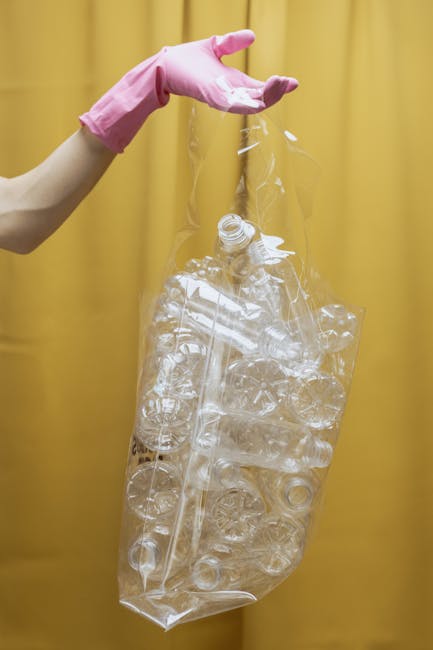 A person's hand wearing a pink rubber glove is holding a large, transparent plastic bag filled with numerous clear plastic bottles and containers, some with visible caps and ridges. The background features a plain yellow curtain or fabric, providing a neutral backdrop that highlights the focus on the waste items. The scene is well-lit, emphasizing the reflective surfaces of the plastic bottles and the crinkled texture of the bag. This image illustrates the handling of recyclable plastic waste, potentially as part of a rubbish removal or recycling process supported by independent waste collection services. The arrangement emphasizes the importance of proper waste segregation and the preparedness for collection or disposal of plastic materials in a domestic or commercial context, aligning with the broader topic of rubbish clearance and alternative waste handling solutions.