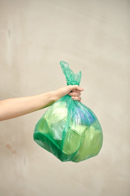 A person's arm extending from the left side of the image holding a green and yellow plastic garbage bag with a knotted top, filled with waste, against a plain, neutral background. The bag appears partly transparent, revealing some contents inside, and is slightly crumpled with visible creases and folds. The scene is well-lit, with soft, even lighting emphasizing the texture of the plastic. This image relates to waste management and rubbish removal services, showcasing a typical collection of household waste prepared for disposal or private waste collection, possibly representing an alternative to local authority rubbish disposal. The focus on the bag and hand highlights the process of rubbish handling, relevant for services offering independent rubbish clearance solutions.