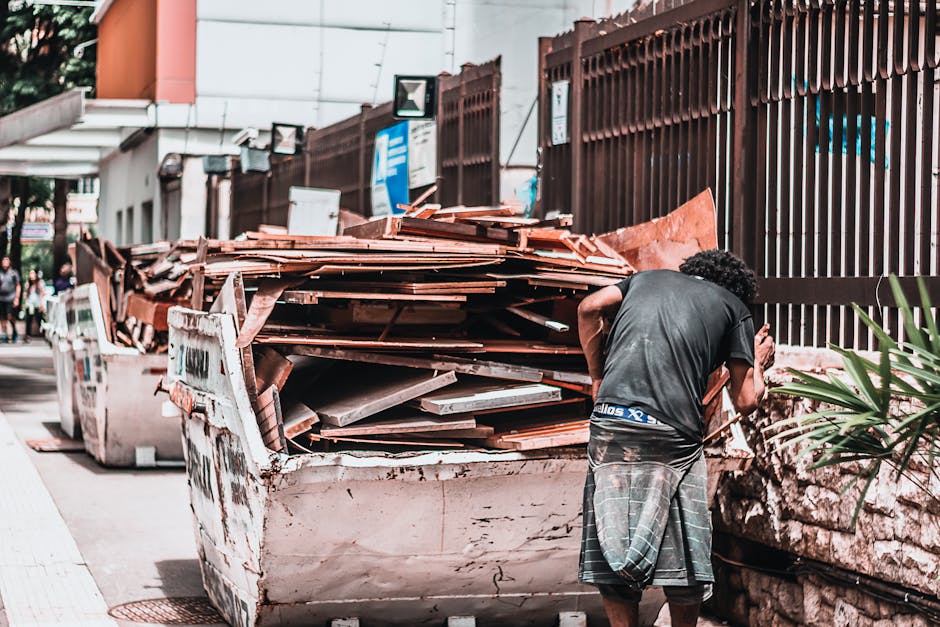 A person with curly dark hair and wearing a black T-shirt and checkered shorts is standing next to a large, rusty white skip filled with a mix of roof tiles, wood planks, and other construction debris. The skip is positioned on a paved pavement beside a tall, dark brown metal fence with vertical bars. In the background, there are signs, a building with white walls, and a few people walking along the sidewalk. The outdoor scene appears to be on a city street, with natural daylight illuminating the area. The surrounding environment suggests a site where private waste disposal or on-site clearance might be taking place, consistent with independent rubbish collection services like those offered by rubbishcollectionkentishtown.co.uk. The materials in the skip, including terracotta tiles and weathered wood, highlight typical waste resulting from renovation or construction activities in an urban area, emphasizing the need for professional waste management solutions.
