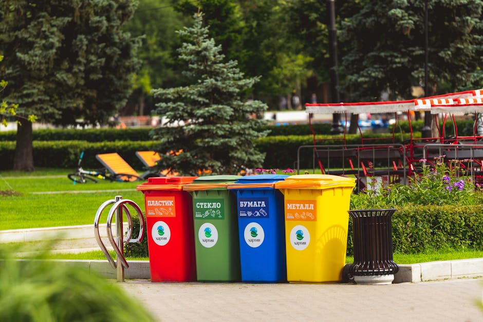 In an outdoor park setting, four separate colour-coded waste bins are lined up on a paved surface next to a grassy area with trees and shrubs. The red bin, labeled with Cyrillic text for plastics, has a textured surface and a hinged lid. The green bin, designated for glass, features an icon of a glass bottle and a hinged lid. The blue bin, intended for paper waste, displays an icon of sheets of paper and also has a hinged lid. The yellow bin, for metal waste, bears an icon of a can and a hinged lid. Behind the bins, a small hedge borders the area, and further back are garden furniture including a swing set and benches, with lush green trees providing a natural backdrop. A black trash receptacle is positioned beside the yellow bin. This scene represents household waste segregation, suitable for private waste collection services rather than municipal disposal, reflecting alternative rubbish handling options often employed in independent rubbish removal in Kentish Town.
